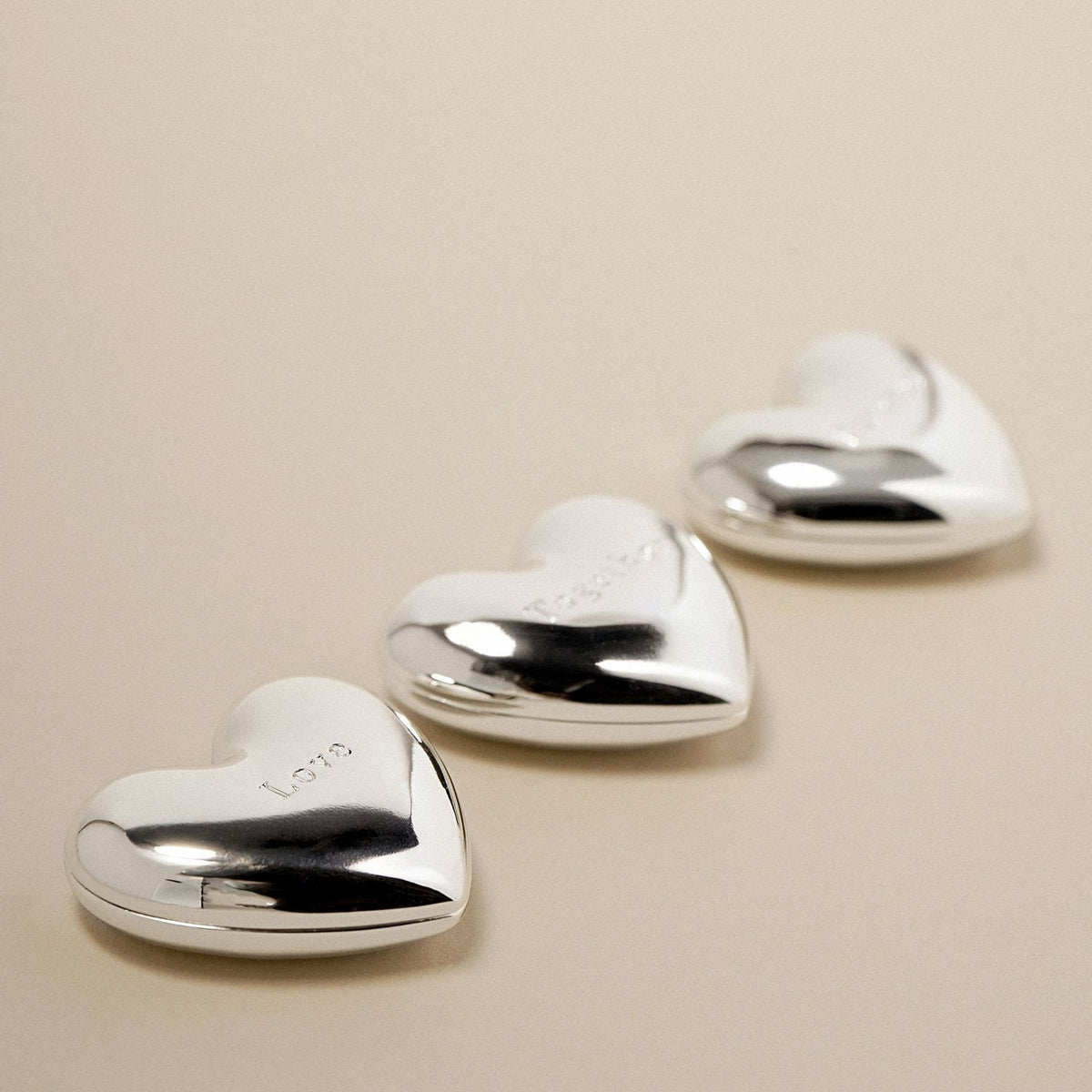 Three silverplated heart charm ornaments with mirror-bright finish, each engraved with "Love", "Forever" and "Together", arranged in a row on a plain beige background.