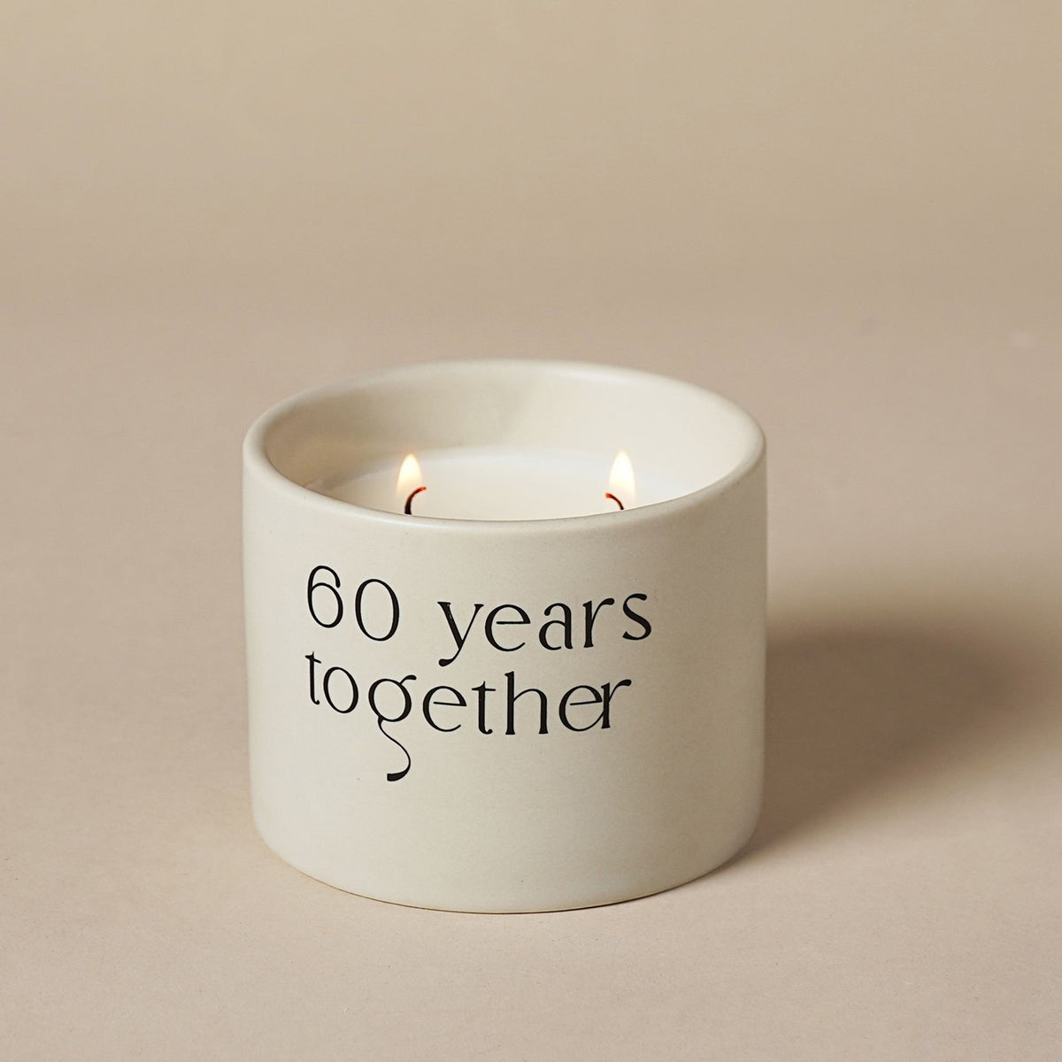 Round white ceramic double wick scented candle with "60 years together" printed in black lettering, two lit flames visible, set against a plain neutral background.