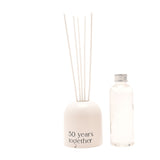 Beige glass reed diffuser with arched, dome-topped holder reading "50 years together", fitted with upright white reeds, beside a clear refill bottle on a white background.