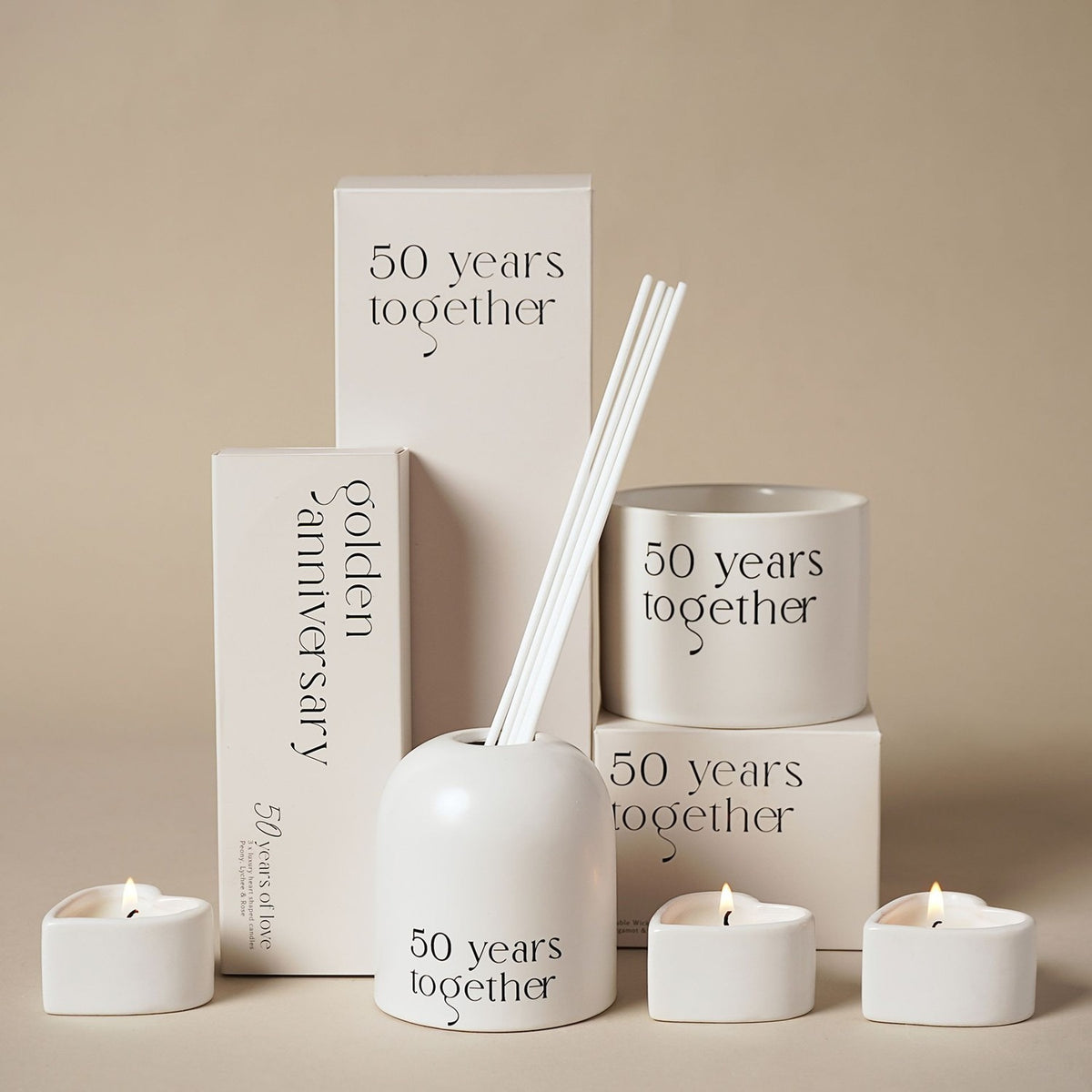 "50 Years Together" glass reed diffuser in beige with arched, dome-topped holder and white reeds, shown with matching tealights and gift boxes.