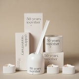 "50 Years Together" glass reed diffuser in beige with arched, dome-topped holder and white reeds, shown with matching tealights and gift boxes.