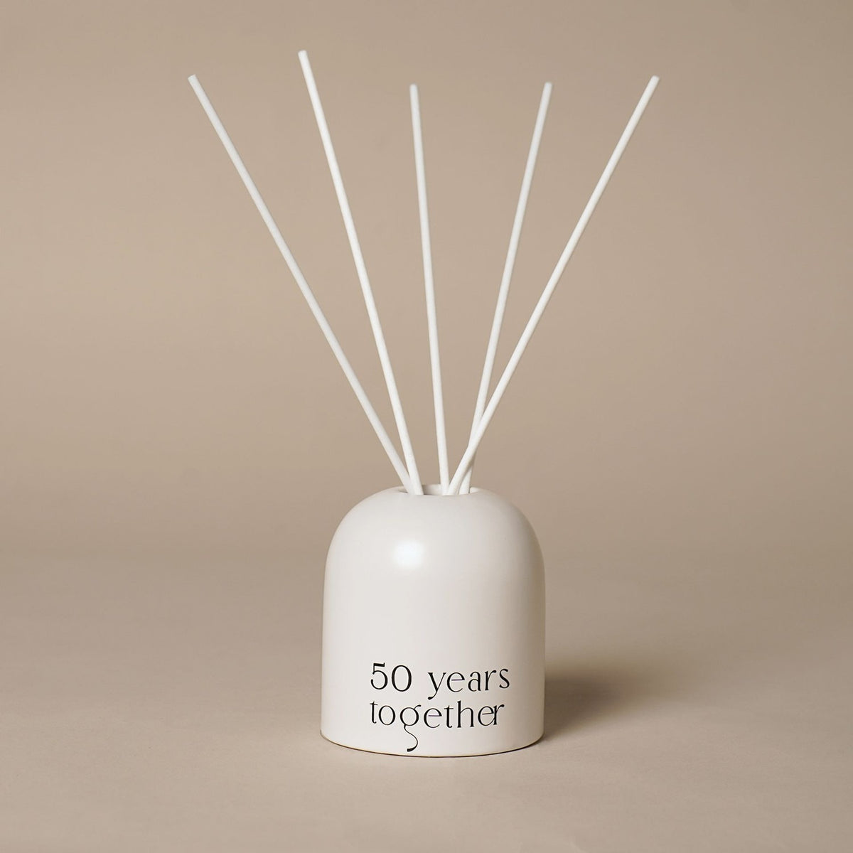 Beige glass reed diffuser with an arched, dome-topped holder, "50 years together" printed in black near the base, and white reeds fanning upward, against a plain neutral background.