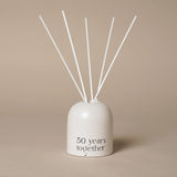 Beige glass reed diffuser with an arched, dome-topped holder, "50 years together" printed in black near the base, and white reeds fanning upward, against a plain neutral background.