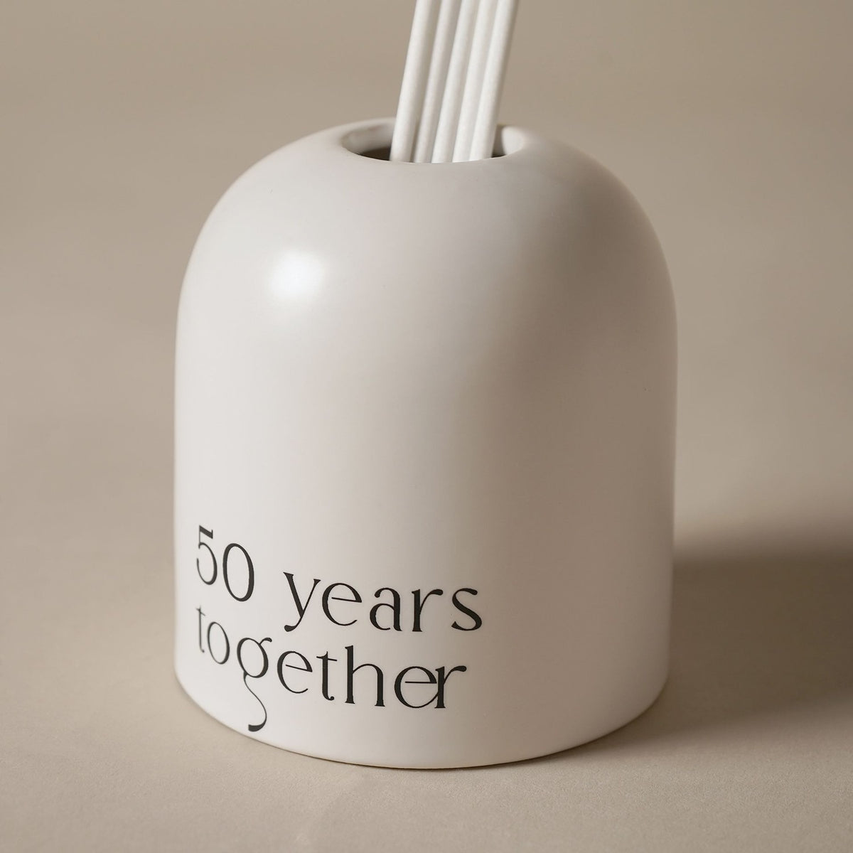 Beige glass reed diffuser with an arched, dome-topped shape, white reeds fanning from the crown, and "50 years together" printed in black lettering on the front, set against a plain light background.