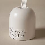 Beige glass reed diffuser with an arched, dome-topped shape, white reeds fanning from the crown, and "50 years together" printed in black lettering on the front, set against a plain light background.