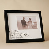 Black wide plastic easel photo frame with "Our Wedding" text and script quote on a white mount, holding a 6 x 4in couple photo, set against a neutral background.