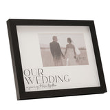 Black plastic easel photo frame with wide woodgrain-effect border, white mount printed "OUR WEDDING, a journey to take together", holding a 6 x 4in wedding photo.