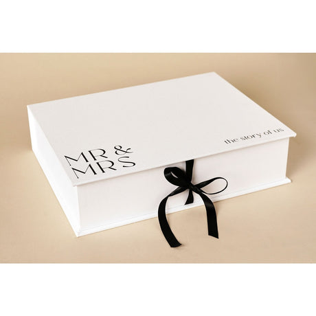 White linen A4 keepsake box with "MR & MRS" and "the story of us" printed on the lid, closed with a black ribbon tie, on a plain background.