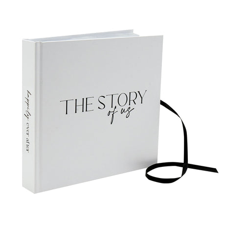 White textured photo album standing upright, with "The Story of us" lettered in black on the cover and a black ribbon tie trailing from the edge, on a plain background.