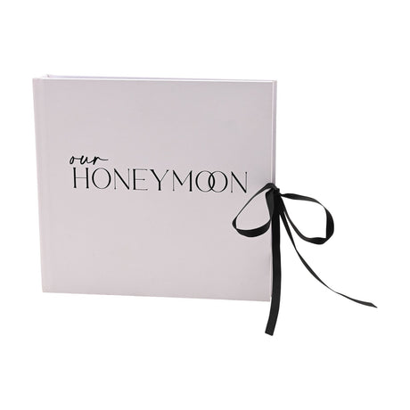 Square "Our Honeymoon" photo album in pale blush pink with black script and capital lettering on the cover, finished with a black ribbon tie bow at the edge, shown on a plain white background.