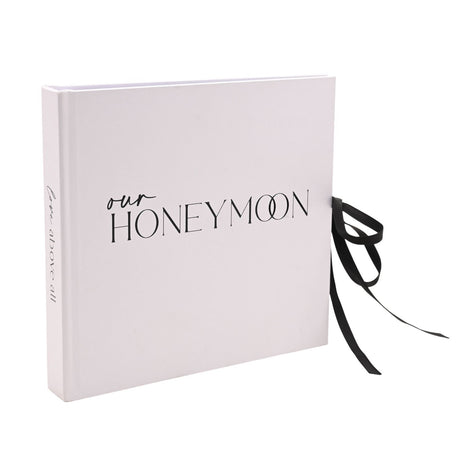 Pale blush pink "Our Honeymoon" photo album with black ribbon tie bow and "love above all" script on the spine, shown against a plain white background.