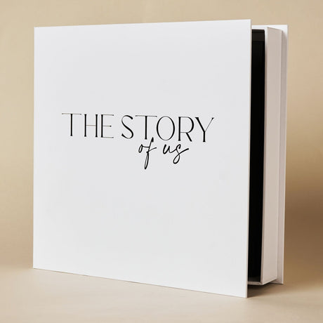 White square photo album, "The Story of Us" printed in black lettering on the cover, shown slightly open to reveal black inner pages, on a plain background.