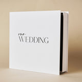 Square white "Our Wedding" photo album in matching box, with 'our' in script and 'WEDDING' in capitals, shown at an angle on a plain background.