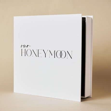 Square white photo album titled "Our Honeymoon" in black lettering, standing upright with visible black inner pages, against a plain beige background.