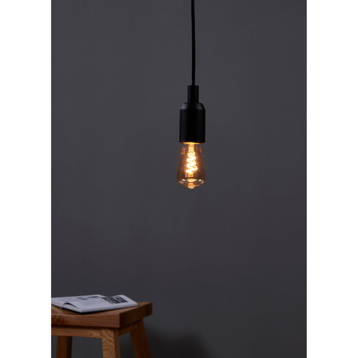 Amber Dimmable LED Bulb E27, tapered vintage-style glass with spiral filament, glowing warm white, shown suspended from a black pendant fitting above a wooden stool against a dark grey wall.
