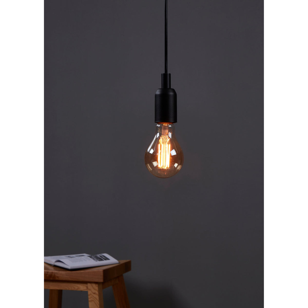 Amber Dimmable LED Bulb E27, A-shape glass with visible filaments, glowing warm white in a black pendant fitting against a dark grey wall.