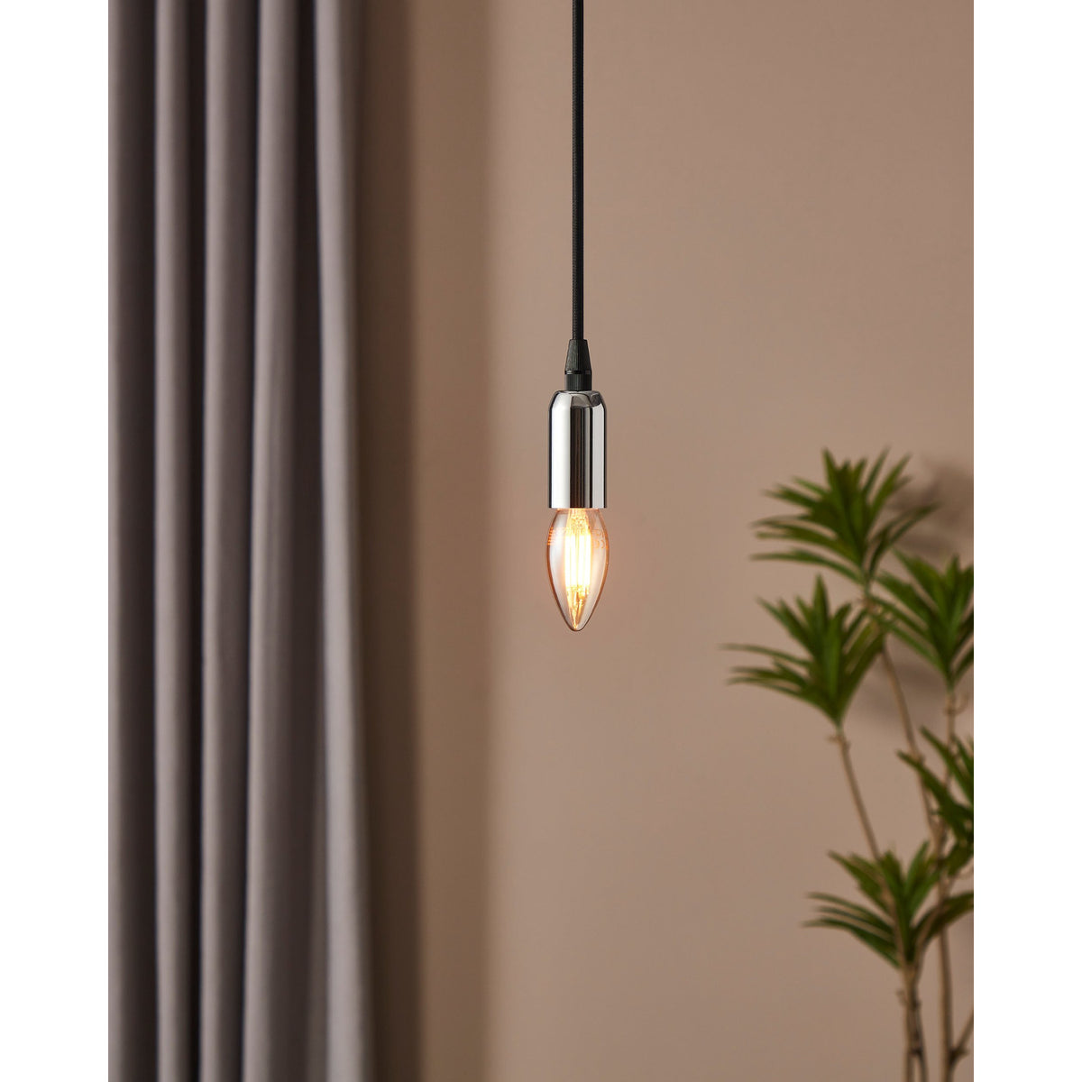 Pendant light with chrome fitting and amber glass LED candle bulb, E14 fitting, glowing warm white, hung against a beige wall near curtains and a plant.