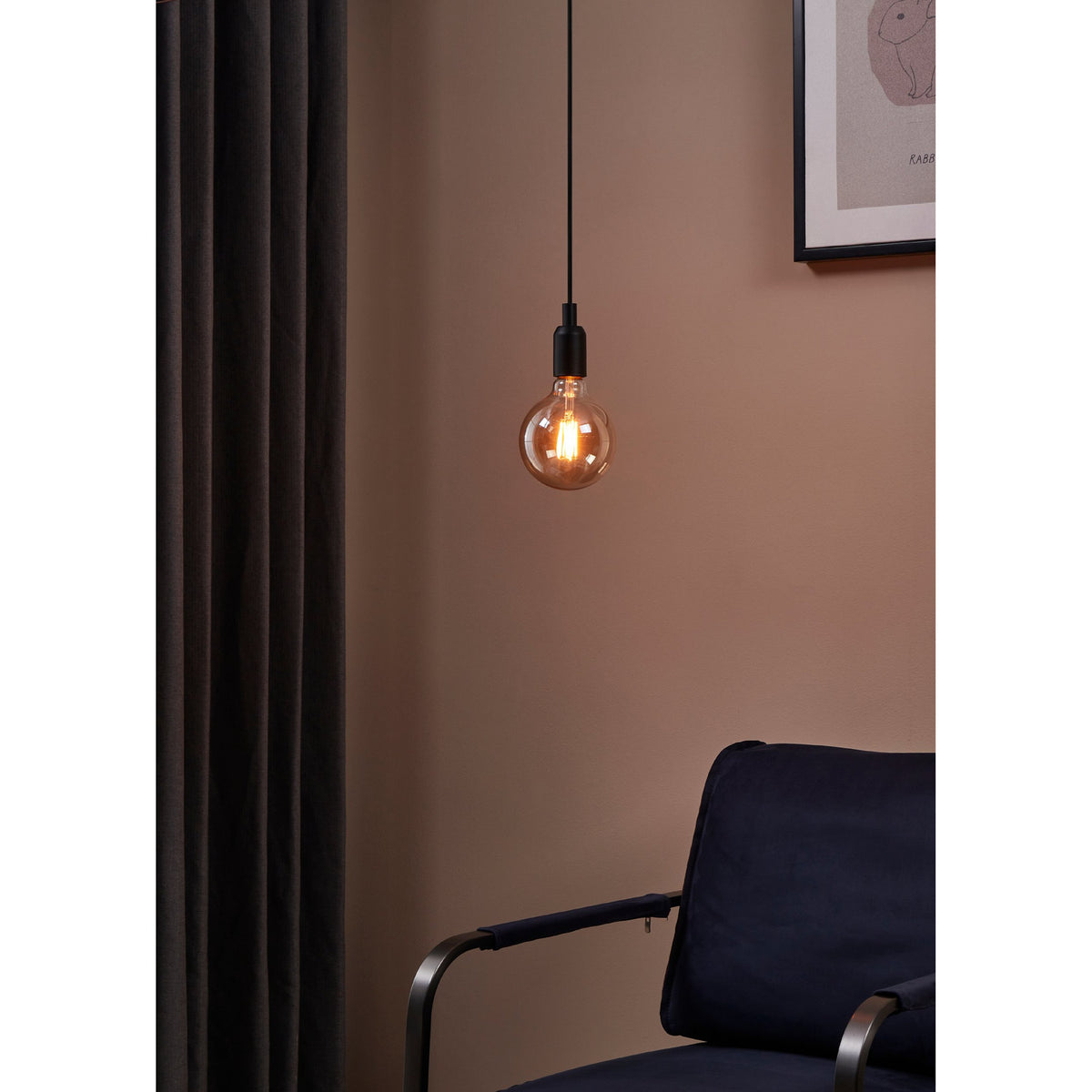 Amber Dimmable LED Globe Bulb E27 G125, visible zigzag filaments and brass cap, hanging lit on a black cord beside a curtain and armchair in a warm-toned room.