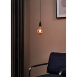 Amber Dimmable LED Globe Bulb E27 G125, visible zigzag filaments and brass cap, hanging lit on a black cord beside a curtain and armchair in a warm-toned room.
