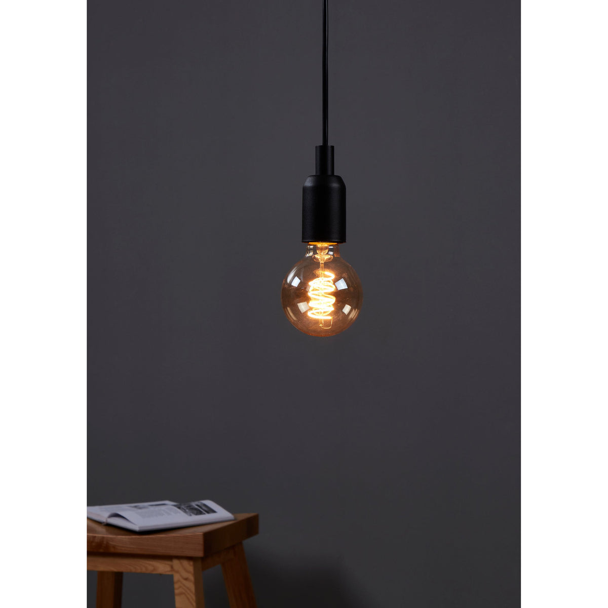 Amber Dimmable LED Globe Bulb E27 G80 4W, lit on a pendant with black cord and fitting, showing its spiral filament, in a room with a wooden stool.