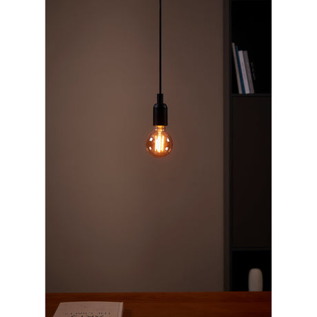 Pendant light fitted with an Amber Dimmable LED Globe Bulb E27 G80, its amber glass and visible filaments glowing warm white above a wooden desk.