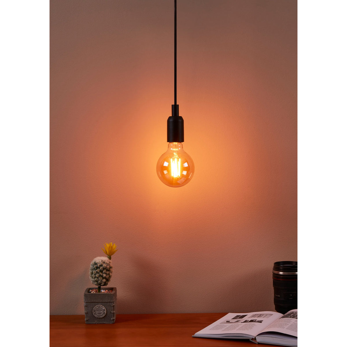 Amber glass G95 LED globe bulb with visible filaments glowing warm white, hung on a black pendant cord above a desk with a cactus, books, and camera lens.