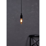 Amber Dimmable LED Tubular Bulb E27 lit warm gold in a black pendant fitting against a dark textured wall, with a black chair visible below.