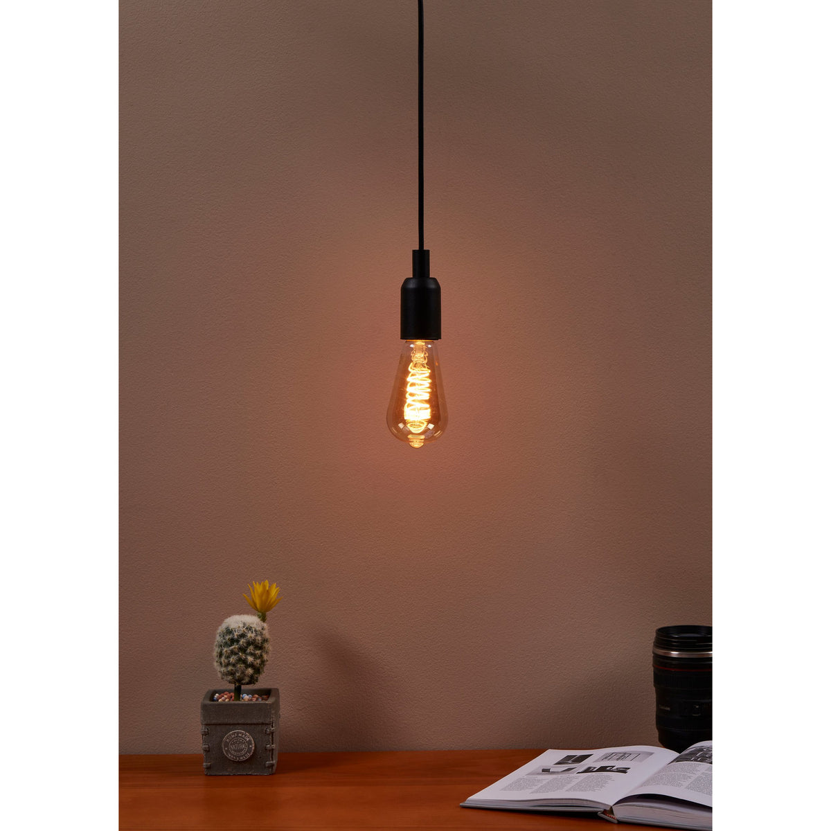 Amber dimmable LED vintage bulb E27 ST64 with spiral filament and teardrop glass shape, shown lit in a black pendant fitting above a desk with a cactus and open book.