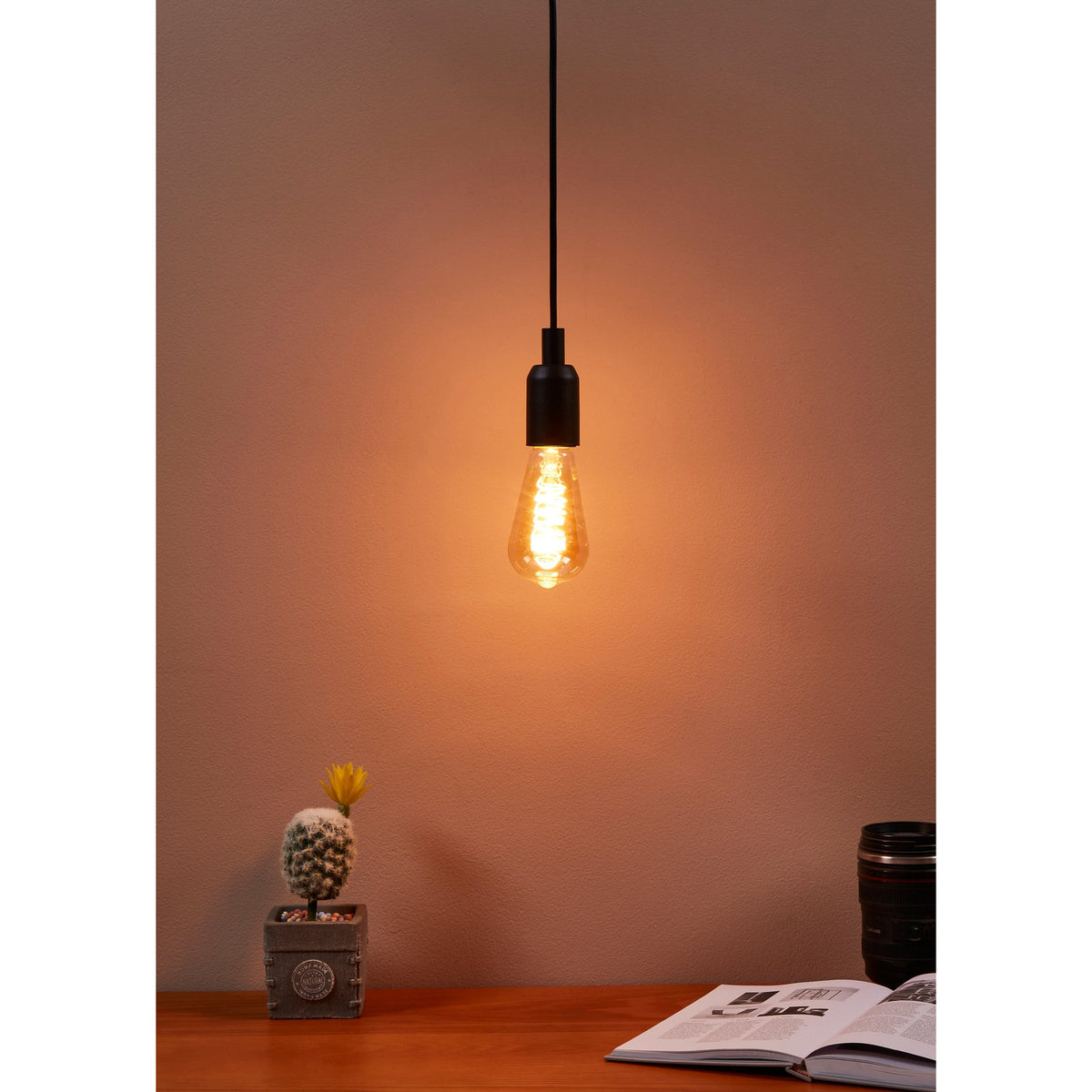 Amber Dimmable LED Vintage Bulb E27 ST64 4W with visible spiral filament, glowing warm white in a black pendant fitting above a desk with a cactus and open book.