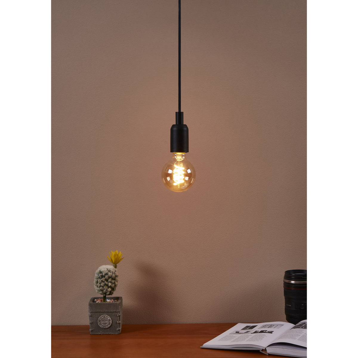 Amber LED Globe Bulb E27 G80 8W warm white, shown lit in a matte black pendant fitting above a desk with a cactus, camera lens, and open book.