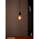 Amber LED Globe Bulb E27 G95 lit warm white, hanging from a black pendant fitting against a muted wall in a room setting.