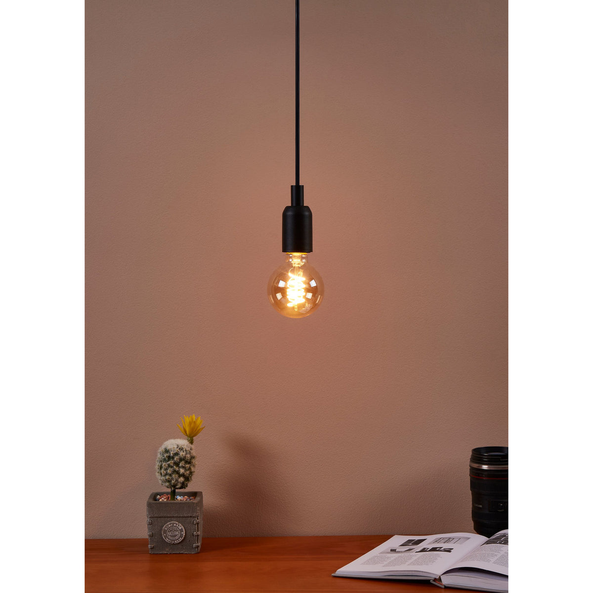 Amber Spiral LED Globe Bulb E27 G80 4W, glowing warm white in a matte black pendant fitting above a desk with a cactus and open book.