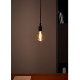 Amber Spiral LED Vintage Bulb E27 ST64, teardrop amber glass with visible spiral filament, glowing warm white, suspended from a black pendant fitting above a wooden desk.