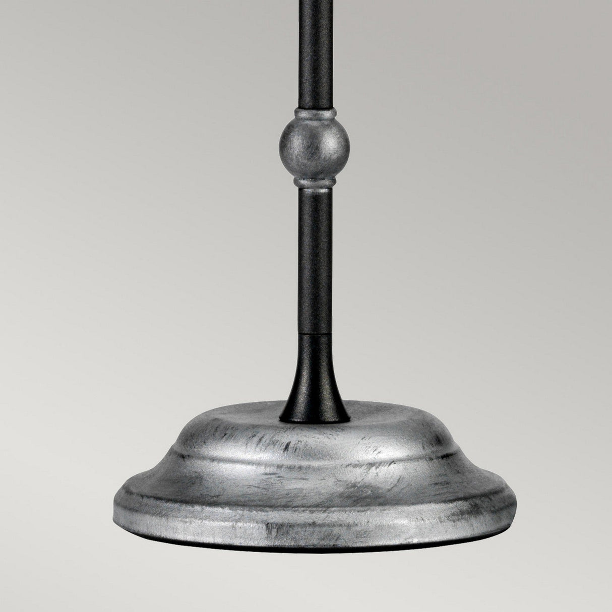 The Amarilli 1 Light Table Lamp - Black With Silver Patina showcases its metal base with a smooth, brushed finish and exquisite handcrafted detailing. A decorative round feature on the pole elevates this lamp to a stunning statement piece against the simple, light gray backdrop.