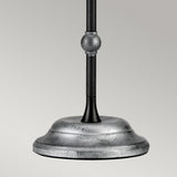 The Amarilli 1 Light Table Lamp - Black With Silver Patina showcases its metal base with a smooth, brushed finish and exquisite handcrafted detailing. A decorative round feature on the pole elevates this lamp to a stunning statement piece against the simple, light gray backdrop.