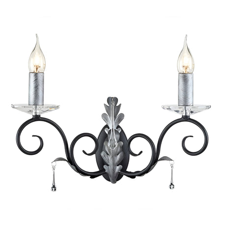 The Amarilli 2 Light Wall Light - Black With Silver Patina from Elstead showcases an elegant and classic design with two candle-like lights beneath clear glass bulb covers. Its ornate black metal frame, accented with silver patina, features scrolls and leaf patterns adorned with hanging crystal droplets.