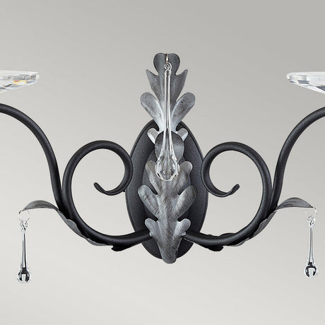 The Amarilli 2 Light Wall Light - Black With Silver Patina adds elegance to any room, with its black and silver patina finish enhancing the ornate scroll-patterned metalwork. Leaf-like details and hanging crystal accents contribute a touch of luxury, akin to the sophistication of an Elstead wall sconce set against a neutral backdrop.