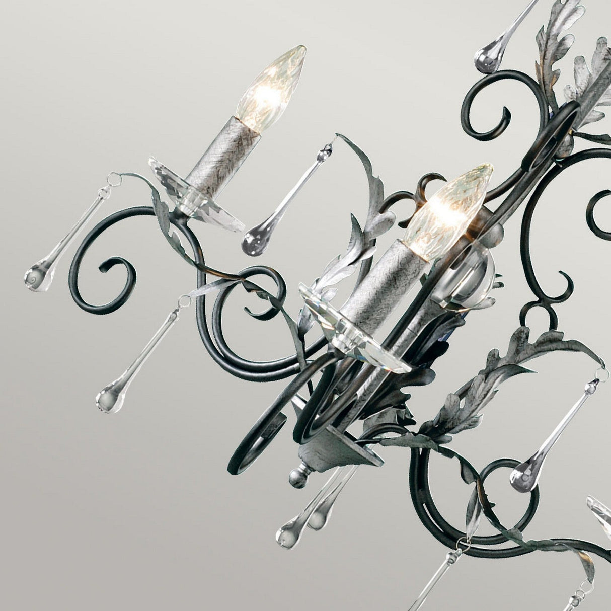 A close-up of the Amarilli 3 Light Chandelier - Black With Silver Patina, with two illuminated bulbs. This handmade Elstead design features intricate metalwork and decorative glass droplets gracefully hanging from the arms against a neutral backdrop.