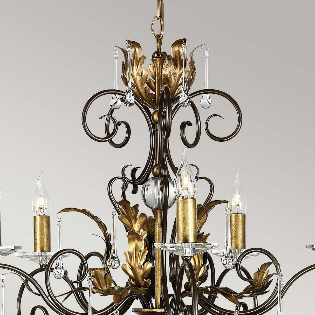 Introducing the Amarilli 5 Light Chandelier by Elstead Lighting, a stunning creation with intricate bronze scrollwork and a gold patina, highlighted by candle-like lights housed in clear glass holders. This ornate design stands elegantly against a light grey background.