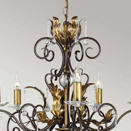 Introducing the Amarilli 5 Light Chandelier by Elstead Lighting, a stunning creation with intricate bronze scrollwork and a gold patina, highlighted by candle-like lights housed in clear glass holders. This ornate design stands elegantly against a light grey background.