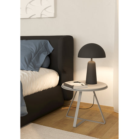 Aranzola Table Lamp in matt black steel with a rounded dome shade on a tapered base, shown on a round bedside table beside a dark upholstered bed.
