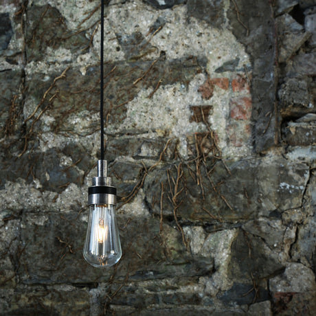 Ardelis Glass Pendant Light IP65, teardrop clear glass shade with silver tiered cap on black cable, lit filament bulb, hung against a stone wall.