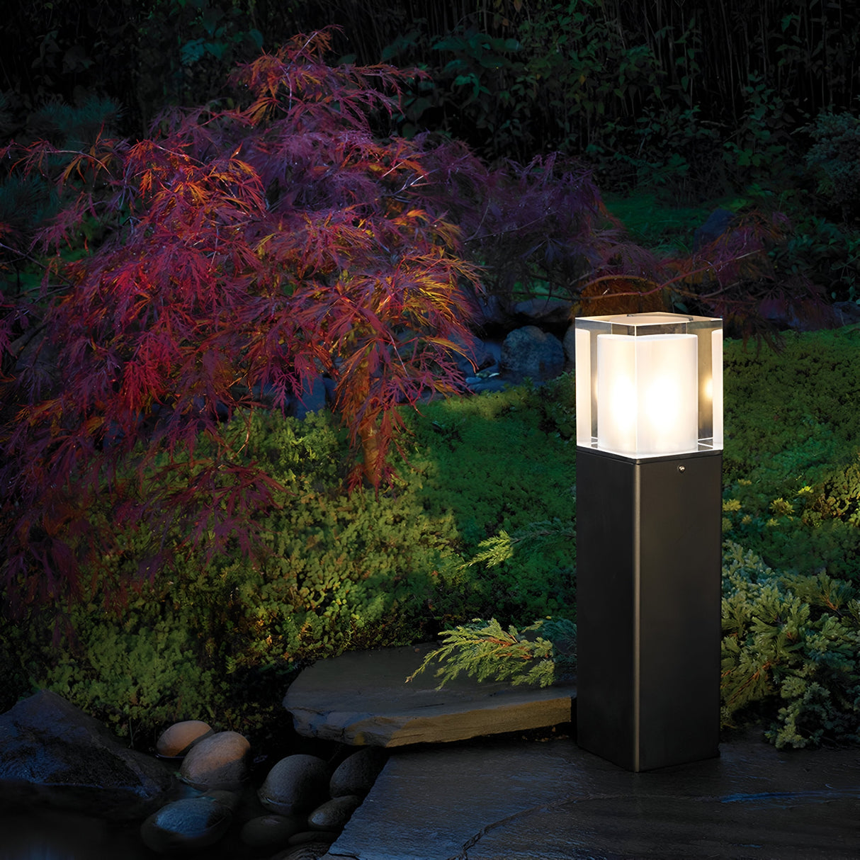 The Arendal 1 Light Medium Bollard in Black bathes a lush garden featuring a vibrant red-leaved shrub and surrounding greenery in gentle light. It stands elegantly on a stone path, casting a warm and inviting glow as dusk falls. Smooth stones are visible in the background, enhancing the charm of this coastal lighting setup.