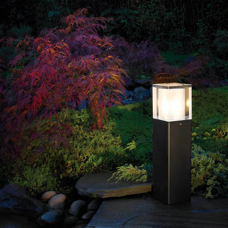 The Arendal 1 Light Medium Bollard in Black bathes a lush garden featuring a vibrant red-leaved shrub and surrounding greenery in gentle light. It stands elegantly on a stone path, casting a warm and inviting glow as dusk falls. Smooth stones are visible in the background, enhancing the charm of this coastal lighting setup.
