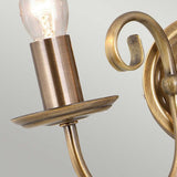 A close-up of the Artisan 2 Light Wall Light in aged brass showcases its artisan craftsmanship with exposed bulbs. The fixture features a curved arm and elegant detailing, highlighted by a decorative scroll design against a pale gray background.