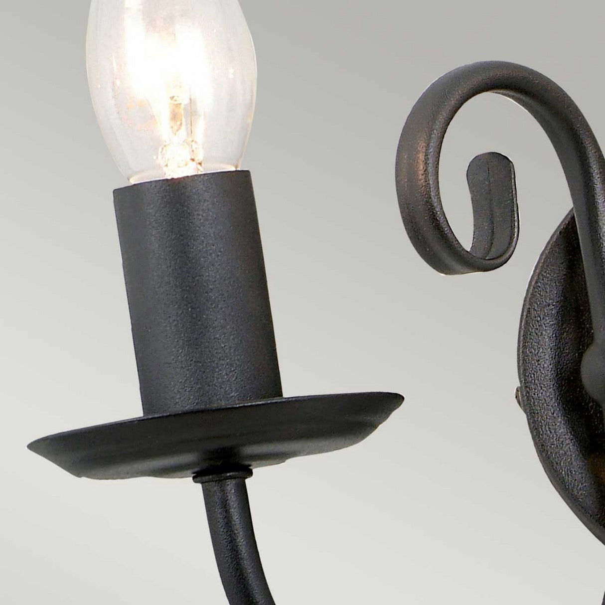 Close-up of the Artisan 2 Light Wall Light - Black, showcasing a black, wrought-iron sconce with a partially visible incandescent light bulb. Featuring artisan craftsmanship, its simple design boasts hand-formed metal scrolls in a sleek black finish against a light gray background.