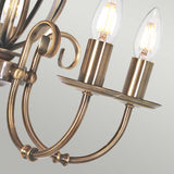 Close-up of the Artisan 5 Light Chandelier in aged brass, showcasing two lit candle-style LED bulbs. This decorative focal point features elegantly curved metal arms and a classic design, set against a neutral background.