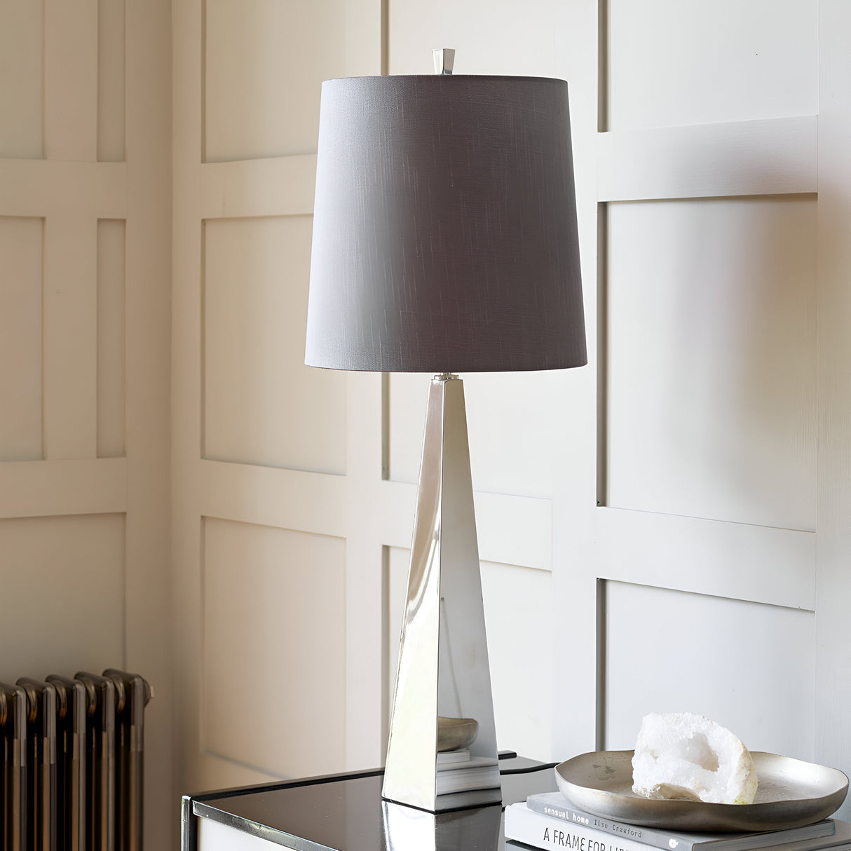 The Ascent Table Lamp With Dark Grey Shade - Polished Nickel elegantly rests on a glass-topped console table. Nearby, a white crystal and a small stack of framed photos add charm, while paneled beige walls provide the perfect backdrop.
