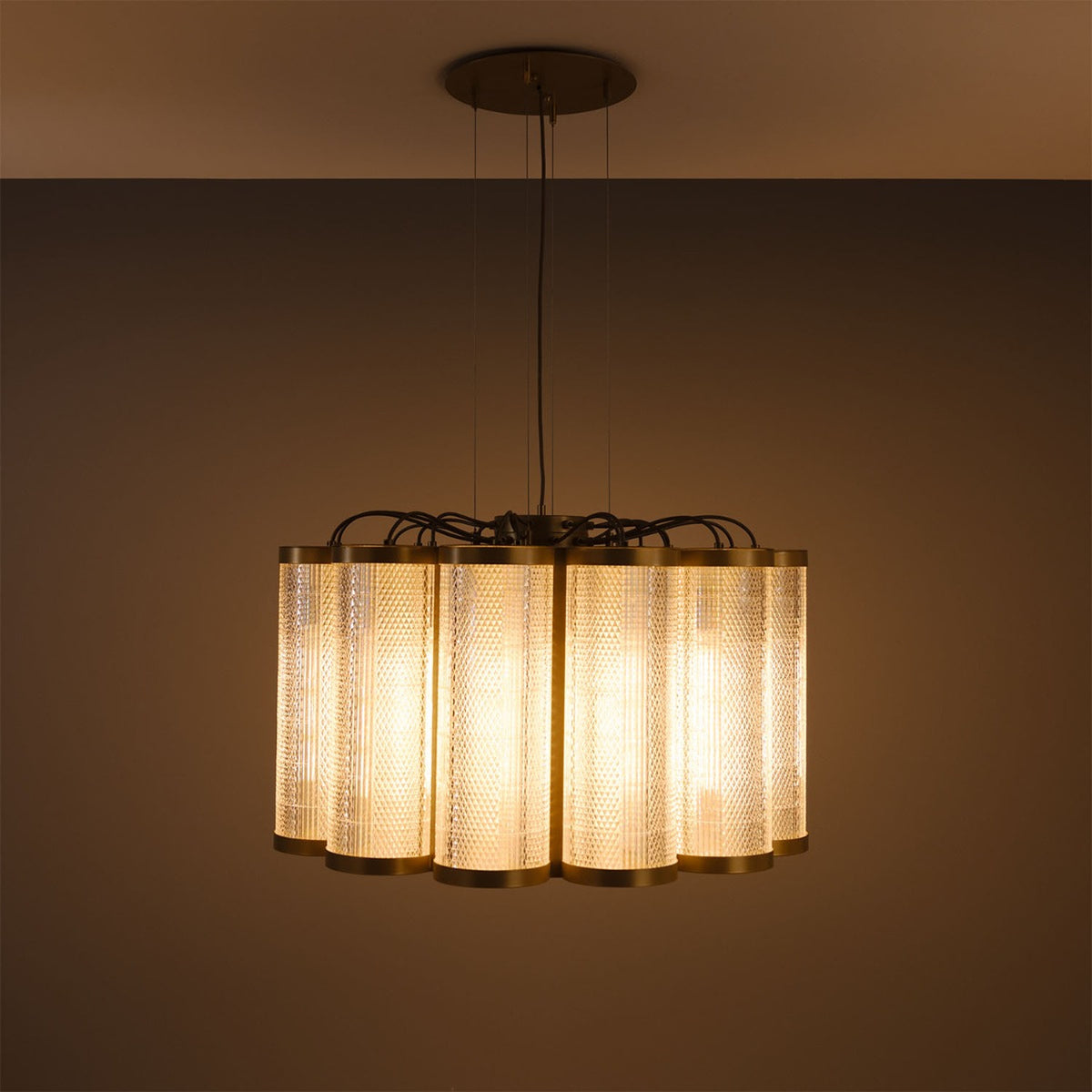 Ashford Acrylic 20 Light Chandelier, a scalloped circle of tall ribbed clear acrylic cylinders with brass rims, suspended from a round ceiling plate, glowing warm against a dark background.