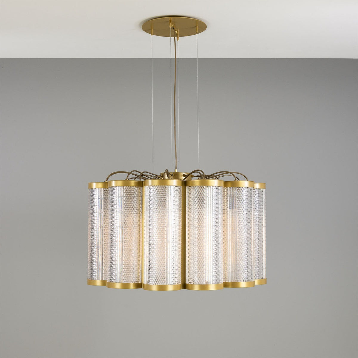 Ashford Acrylic 20 Light Chandelier, a scalloped circle of ribbed clear acrylic cylinders with brass rims, hung from a round brass ceiling plate against a grey wall.